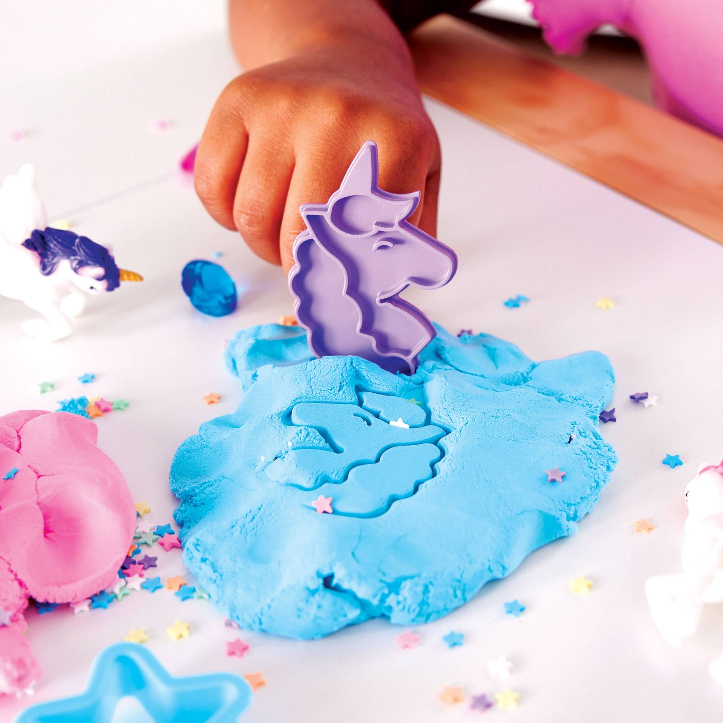 Child's hand using a unicorn-shaped mold in blue play dough with colorful sprinkles on a white surface.