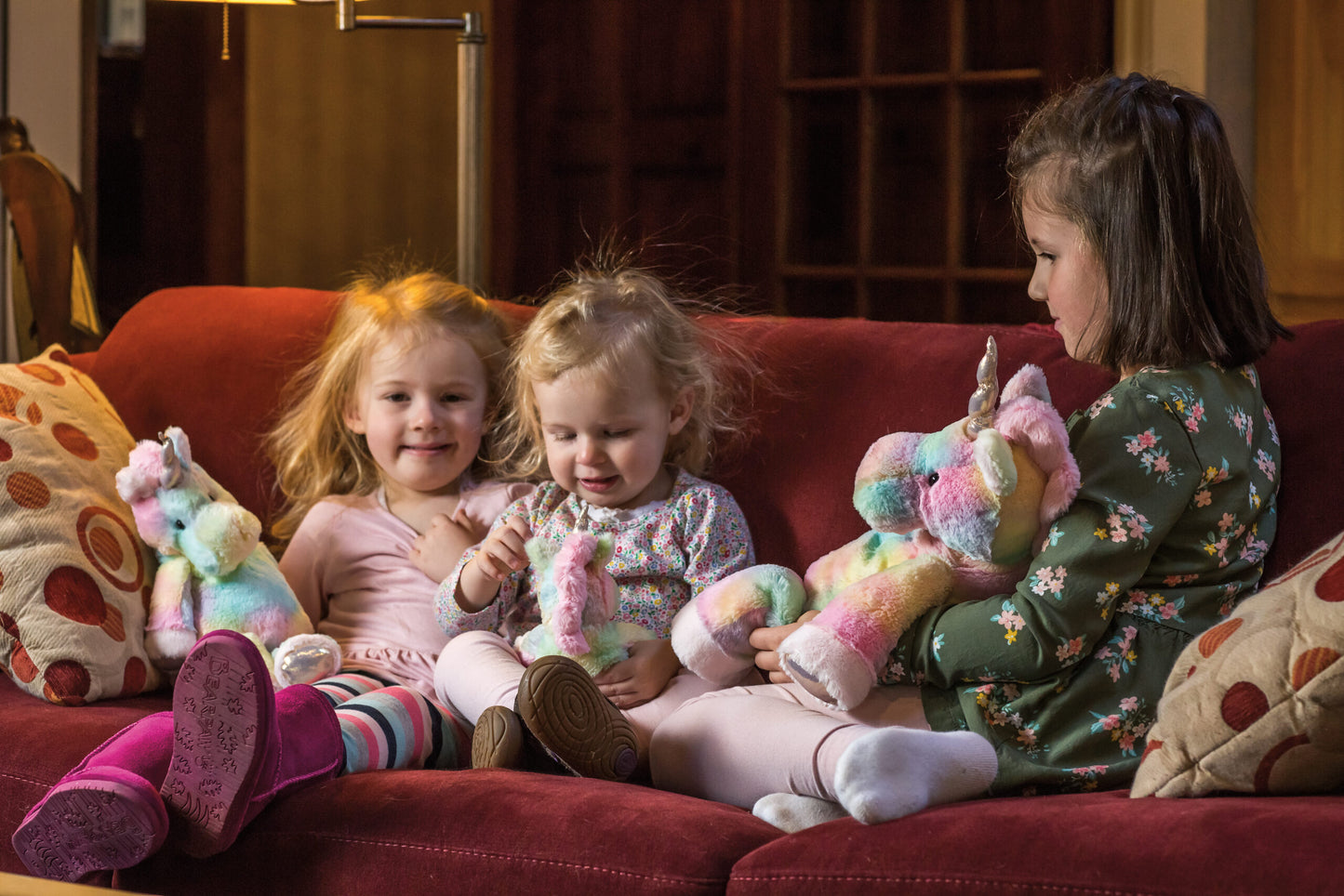 Three children sitting on a red couch with colorful stuffed toys.