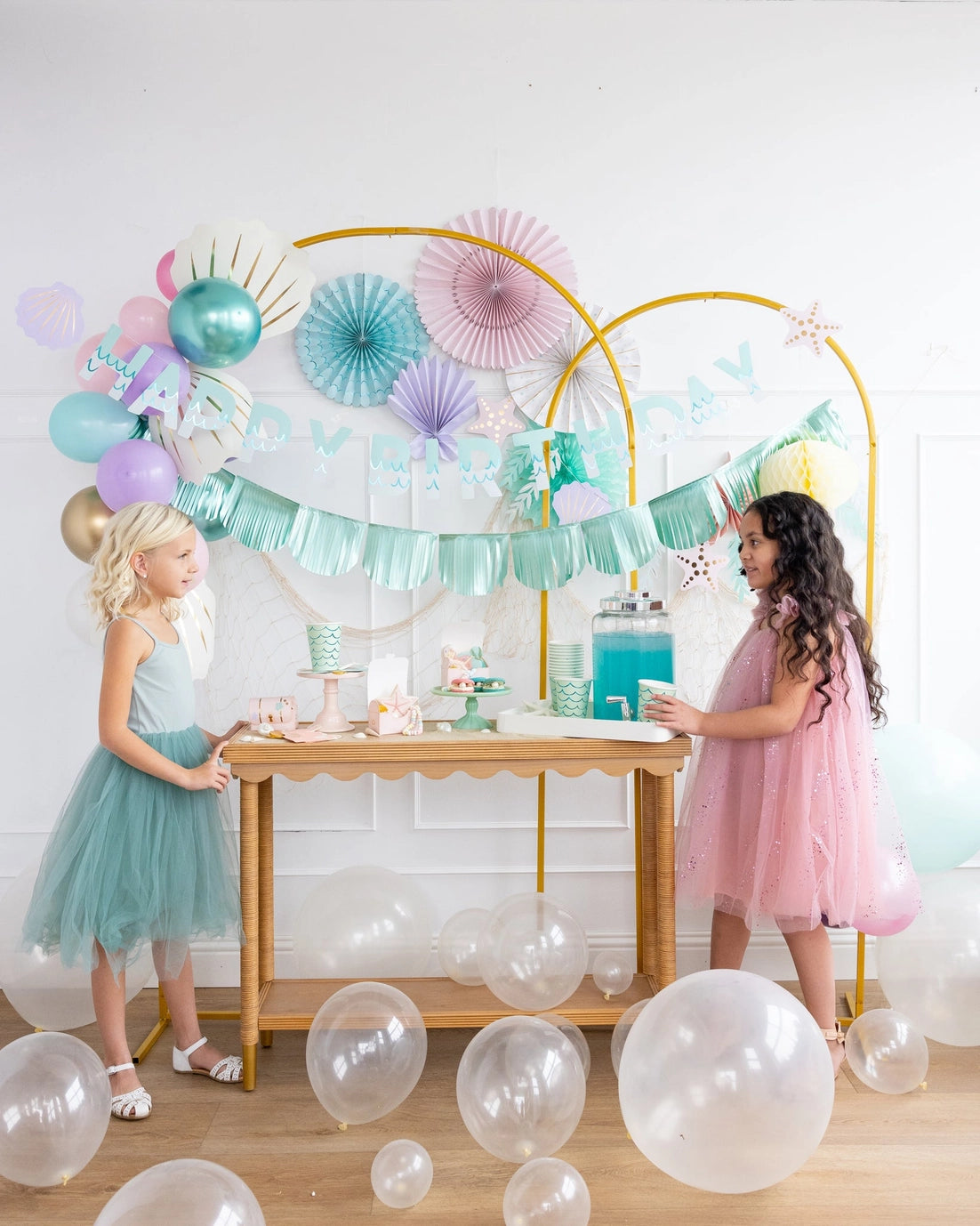 Two young girls in pastel dresses standing behind a table with a decorative arch and balloons.