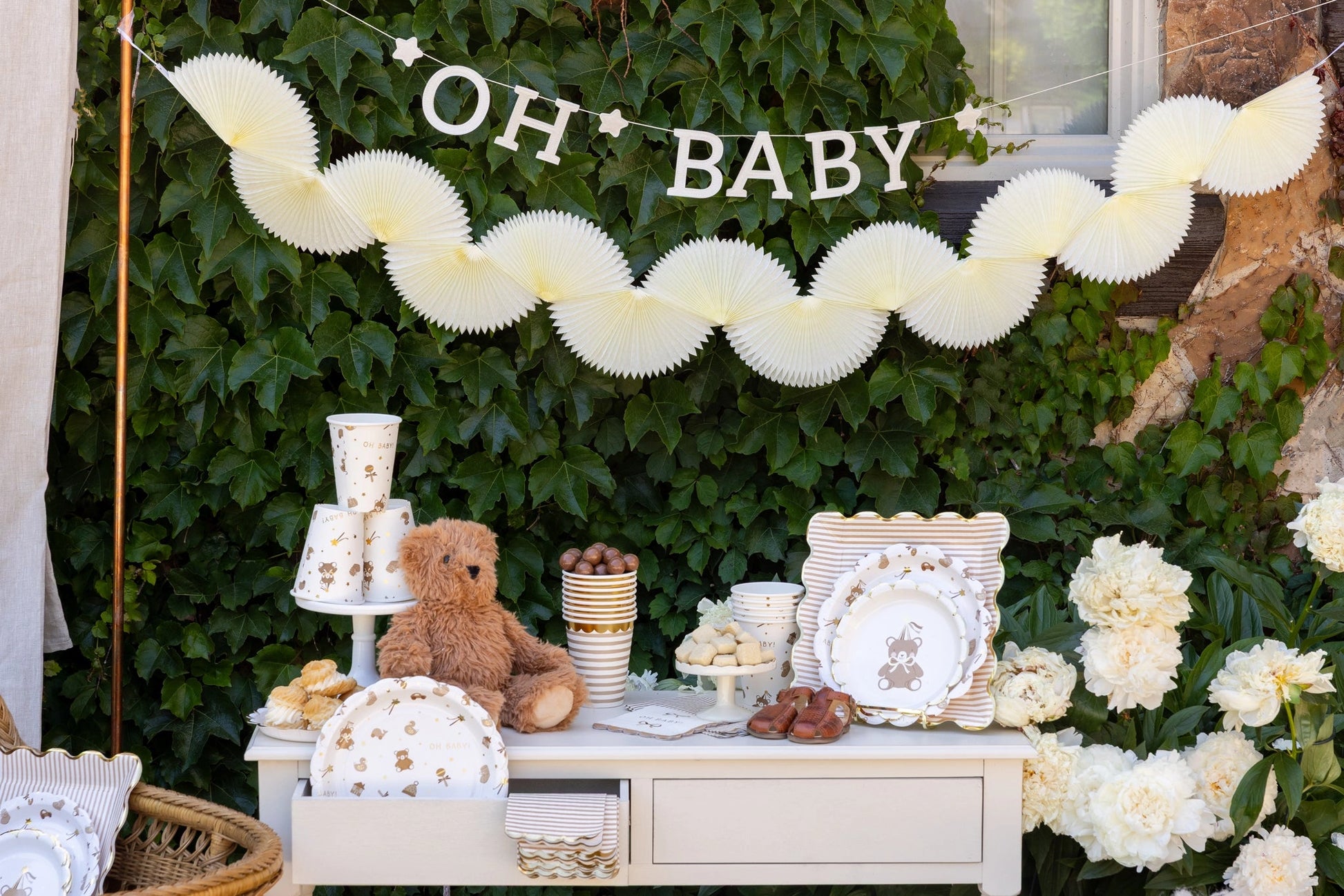 Baby shower setup with 'OH BABY' banner, teddy bear, and decorative items against a green leafy background.