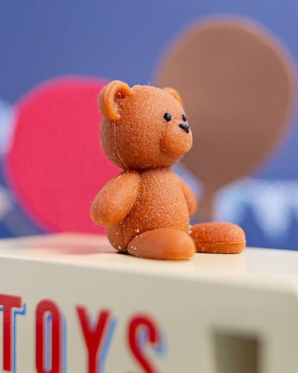 Small brown teddy bear on a block with 'TOYS' letters against a blue background with speech bubbles.