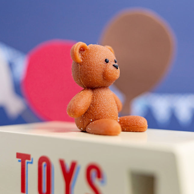 Small brown teddy bear on a block with 'TOYS' letters against a blue background with speech bubbles.