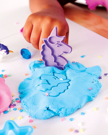 Child's hand using a unicorn-shaped mold in blue play dough with colorful sprinkles on a white surface.