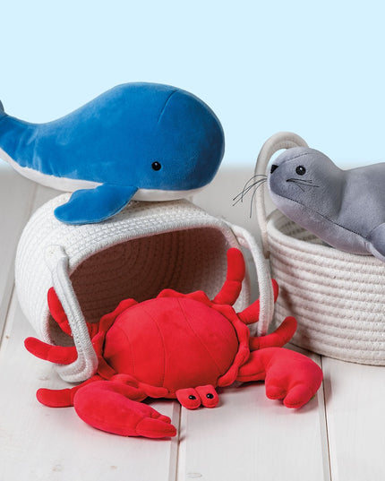 Plush toys including a blue whale, red crab, and gray seal in white baskets on a light wooden floor.