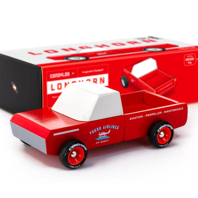 Red toy truck with white top, next to its packaging on a white background