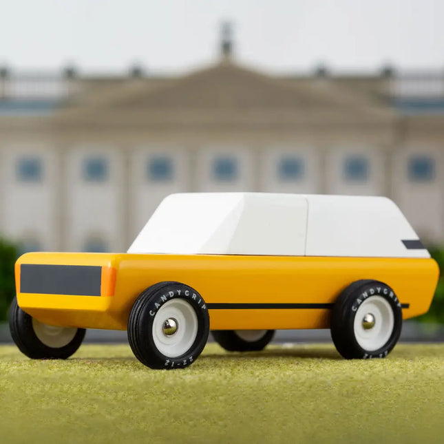 Yellow toy car with a white top on a grassy surface with a blurred building in the background