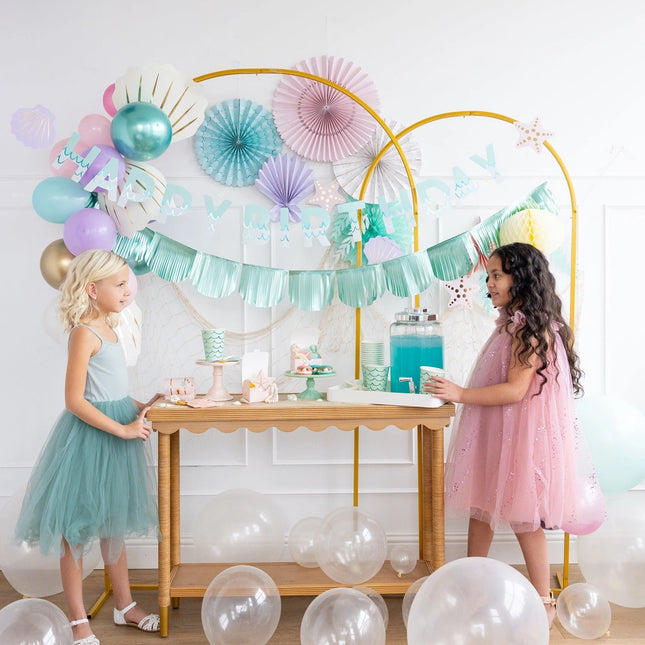Two young girls in pastel dresses standing behind a table with a decorative arch and balloons.