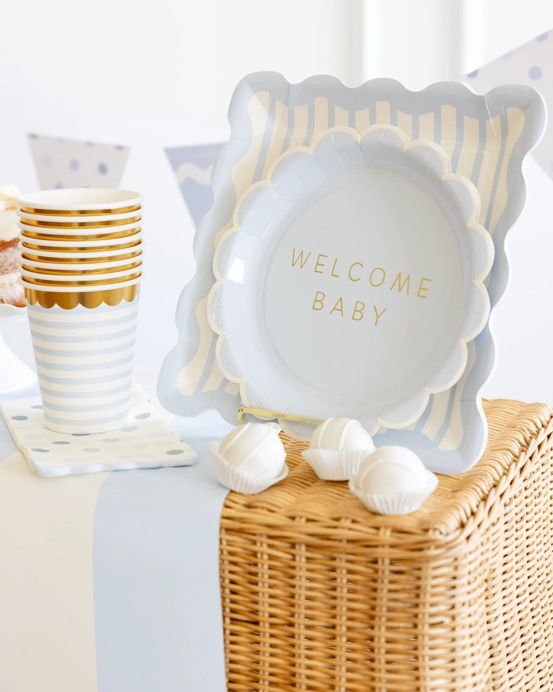 Plates with 'Welcome Baby' text on a white surface and this plate behind it with wicker basket and gold cups.