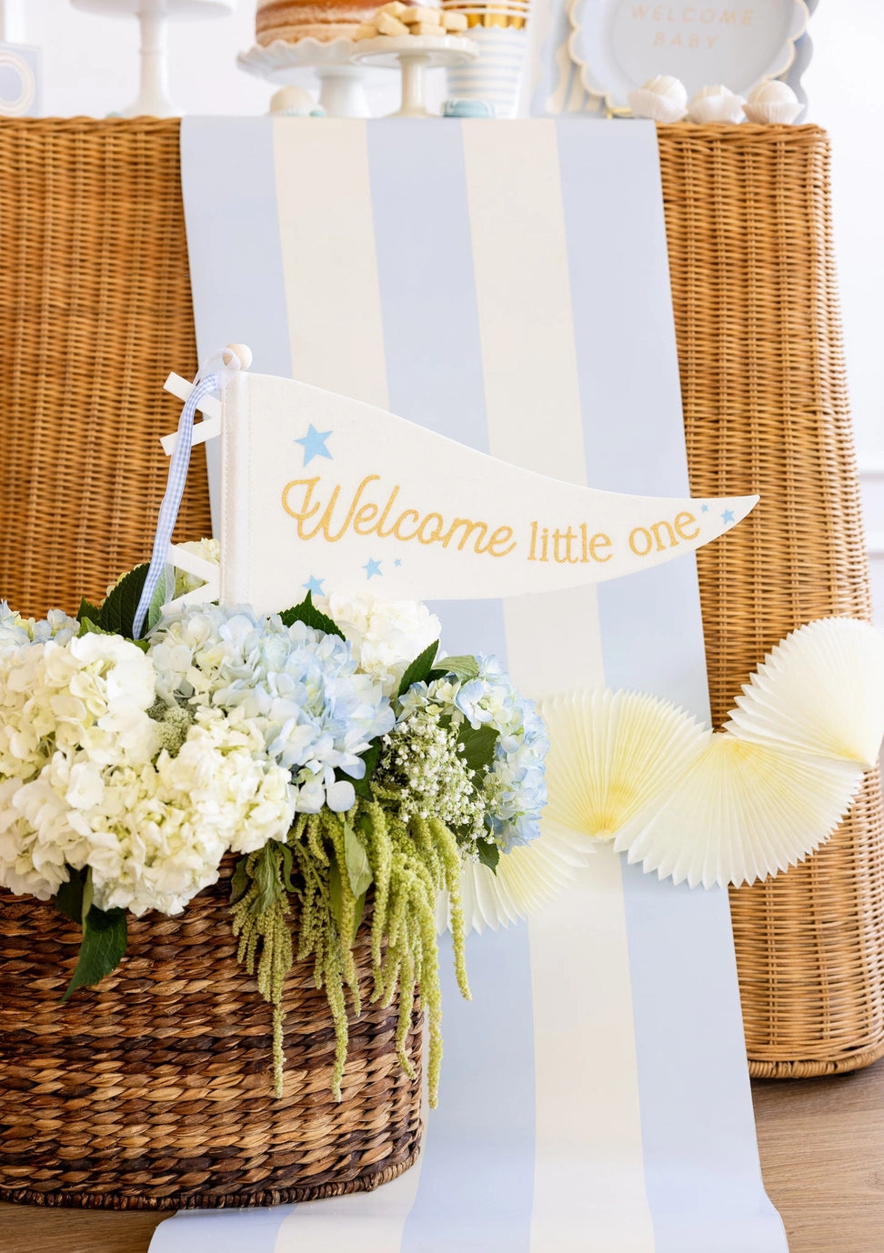 Decorative setup with flowers, a basket, and a 'Welcome little one' sign on a light blue background.