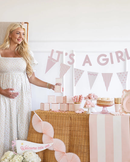 Pregnant woman standing next to a baby shower setup with 'It's a Girl' banner.