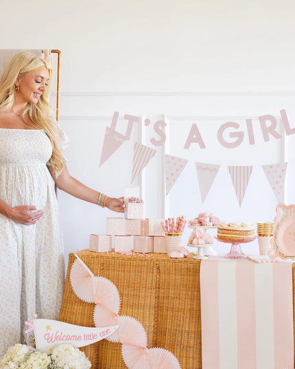 Pregnant woman in a white dress standing next to a 'It's a Girl' themed baby shower table.
