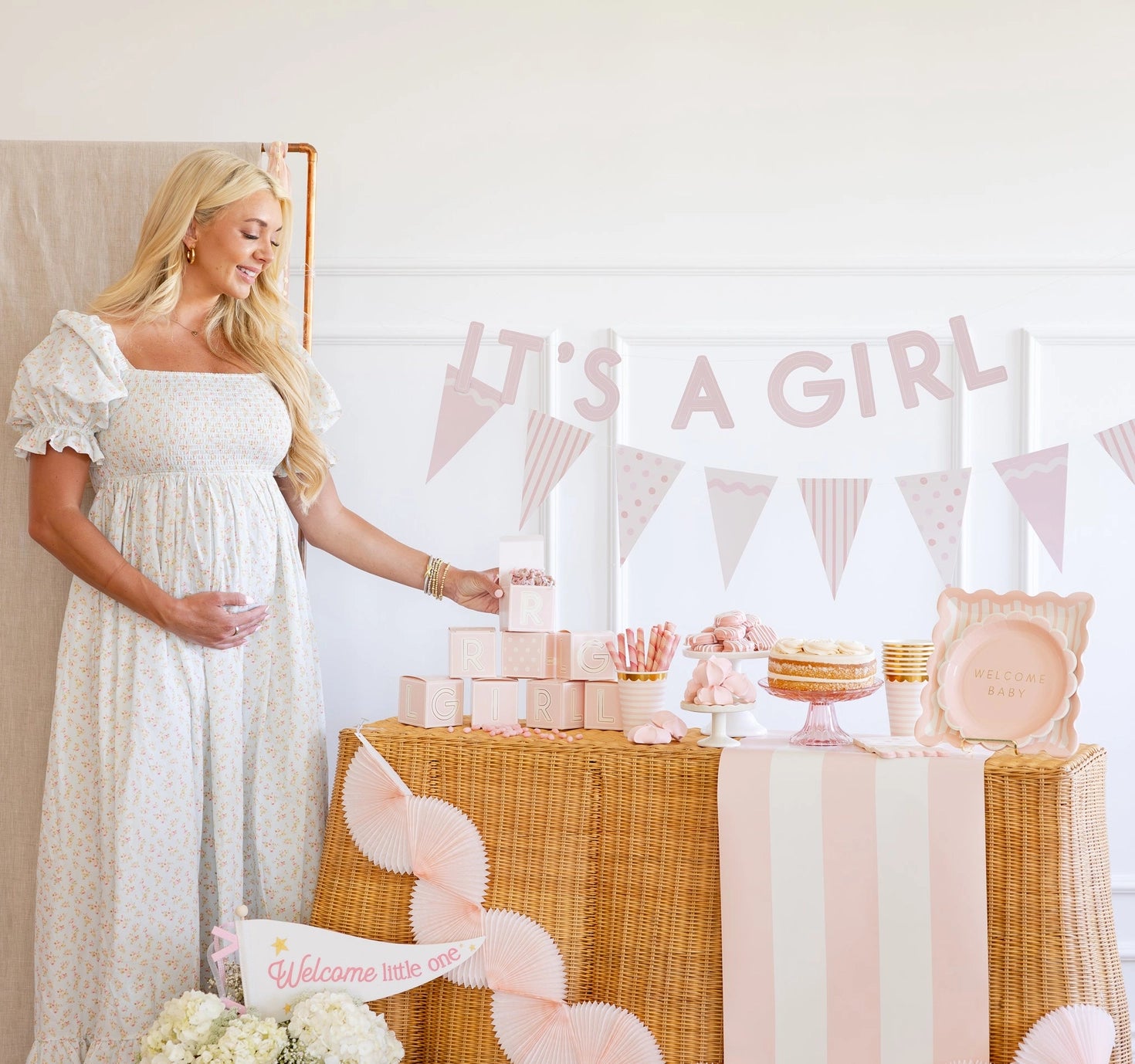 Pregnant woman in a white dress standing next to a 'It's a Girl' themed baby shower table.